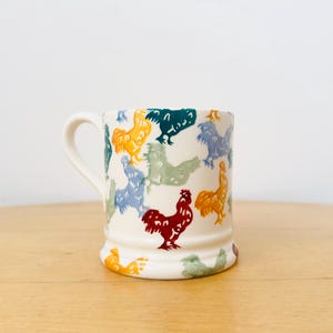 May include: A cream-coloured ceramic mug featuring a repeating pattern of roosters in various colours including red, yellow, blue, green, and teal. The mug has a cylindrical body with a slightly flared base and a curved handle on the left side.