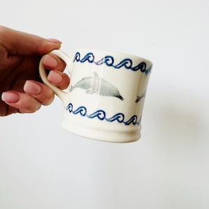 Może przedstawiać: Kremowy ceramiczny kubek z niebieskim wzorem fal i szarą ilustracją delfina. Kubek ma zakrzywiony uchwyt. Wzór powtarza się wokół korpusu kubka. Kubek jest trzymany w dłoni.