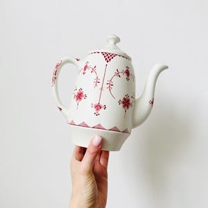 Puede incluir: Una tetera de cerámica blanca con tapa, asa y boquilla. La tetera está decorada con un estampado floral rojo. La tetera está sujeta por una persona. Es un artículo de menaje.