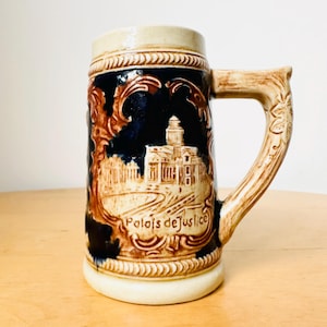 Op de afbeelding: Een bruine en witte keramische bierkan met een blauw en bruin ontwerp. De kan heeft een gebouw met de tekst "Palais de Justice" aan de voorkant.