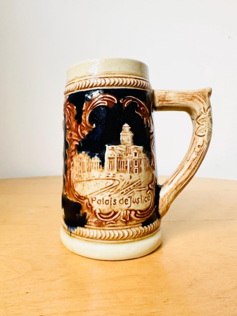 Op de afbeelding: Een bruine en witte keramische bierkan met een blauw en bruin ontwerp. De kan heeft een gebouw met de tekst "Palais de Justice" aan de voorkant.