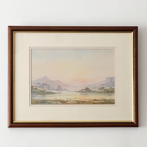 May include: A framed watercolour landscape painting featuring a calm lake reflecting a soft sunrise, surrounded by distant mountains and rolling hills. The artwork is encased in a dark wood frame with a gold inner trim and a cream-coloured mat board.