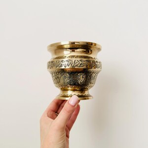 Può includere: Un piccolo vaso in ottone ornato, con un bordo largo e una base a piedistallo. Il vaso presenta intricati motivi floreali e di foglie incisi attorno al corpo. L'ottone lucido ha una calda tonalità dorata, che riflette la luce. Il vaso è tenuto da una mano su sfondo bianco.