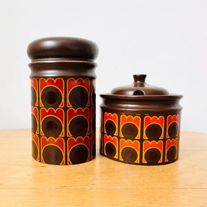 May include: Two ceramic kitchen canisters. One is tall and cylindrical, the other is shorter with a lid. Both are brown with a repeating pattern of orange, red, and brown shapes. The canisters are on a light-coloured wooden surface.