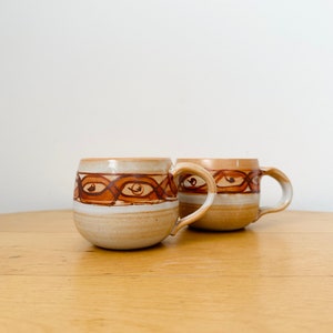 Puede incluir: Dos tazas de cerámica marrón con una franja blanca y un diseño geométrico marrón. Las tazas tienen una forma redondeada y un asa a cada lado.