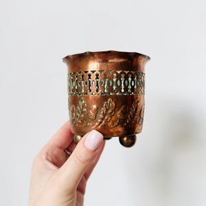 May include: A copper-toned metal planter with an ornate design. The planter features a detailed pattern of leaves and acorns around the base and a pierced band around the middle. It is supported by three small, round feet.