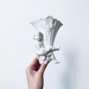 Könnte beinhalten: Weiße Keramikvase mit Cherub-Design. Die Vase hat eine geriffelte Oberseite und einen geformten Cherub, der eine Traube hält. Ein dekoratives Stück für die Inneneinrichtung.