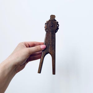 May include: A dark brown wooden nutcracker with detailed carvings. The top has a circular design with radiating patterns, and the handle features a geometric pattern. The nutcracker is held against a white background.