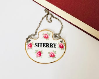 May include: A white ceramic Sherry wine label with a gold trim and a silver chain. The label is decorated with pink roses and the word "SHERRY" in black letters. A red book is in the background.