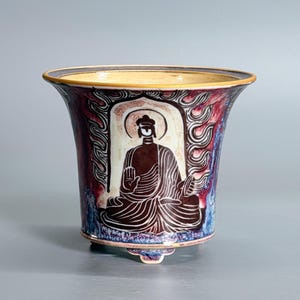 May include: A ceramic planter with a flared rim and three small feet. The exterior features a dark brown, stylized seated figure with a halo, surrounded by intricate flame-like patterns in shades of red, blue, and white against a cream background.
