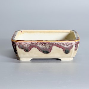 May include: A rectangular ceramic bonsai pot with a cream-colored base and a decorative drip glaze pattern in shades of deep burgundy, dark purple, and black along the upper rim. The pot features a smooth interior and four small feet.