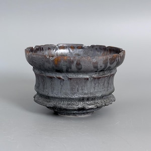 May include: A dark, metallic-toned ceramic bowl with a wide, flared rim and a textured, rugged exterior surface. The glaze features mottled shades of charcoal, black, and deep bronze, creating a weathered appearance with a distinct, uneven base.