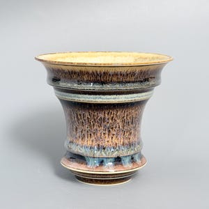 Może przedstawiać: Ceramiczny wazon z szerokim, rozszerzającym się brzegiem i zwężającą się podstawą. Zewnętrzna powierzchnia posiada złożoną glazurę z warstwami głębokiego brązu, beżu i kremu, podkreśloną pionowymi zaciekami oraz subtelnym niebiesko-szarym pasem.