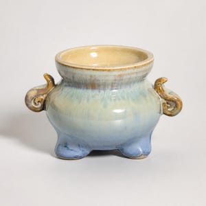 Puede incluir: Un quemador de incienso de cerámica con cuerpo redondeado y esmalte en tonos azul claro y crema. El recipiente cuenta con tres patas cortas, una amplia abertura circular y dos asas decorativas curvadas en los laterales de color marrón.