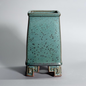 Op de afbeelding: Een vierkante keramische vaas met een blauwgroen glazuur en een donkere gespikkelde textuur. De vaas heeft een taps toelopend lichaam en staat op vier korte, stevige voetjes, elk versierd met een verhoogd, geometrisch Grieks sleutelpatroon.