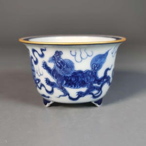 Peut inclure: Un bol en porcelaine blanche orné de motifs peints à la main en bleu sous glaçure représentant des lions mythiques et des nuages. Le bol présente un bord évasé avec un fin liseré doré et repose sur trois petits pieds courts et arrondis.