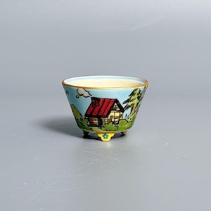 May include: A small ceramic bowl with a light blue exterior featuring a hand-painted scene of a cabin with a red roof, green trees, and hills. The bowl has a cream-colored interior, a gold-toned rim, and three small yellow feet with blue flower details.