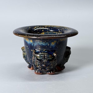 May include: A dark blue ceramic planter featuring a wide, flared rim and a glossy finish with gold-toned speckles. The exterior is decorated with three sculpted, relief-style animal figures that serve as feet, supporting the rounded vessel body.