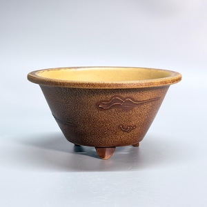 Puede incluir: Un cuenco de cerámica redondo y cónico con un exterior texturizado de color marrón moteado y un interior liso de color crema claro. El cuenco presenta un borde ancho y plano y se apoya sobre tres pies pequeños y robustos con detalles en relieve.