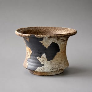 May include: A ceramic bowl with a wide, flared rim and a footed base. The exterior features a contrasting design of matte black sections interspersed with textured, mottled beige and brown patches, creating a rugged, uneven surface appearance.