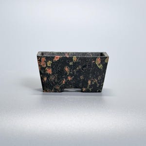 May include: A rectangular bonsai pot with a speckled design. The pot is primarily black with orange and yellow flecks. It has a slightly tapered shape and small feet. The pot is designed for holding small plants.