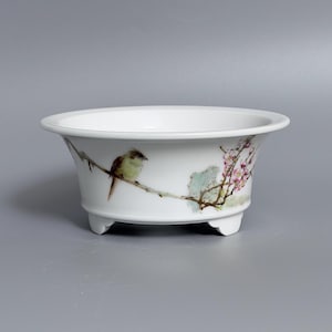 May include: A white ceramic bowl with a flared rim and three small feet. The exterior features a hand-painted illustration of a small brown bird perched on a thin branch with delicate pink blossoms and green leaves extending across the side.