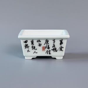 Könnte beinhalten: Ein rechteckiger weißer Keramik-Pflanztopf mit ausgestelltem Rand und vier kleinen Füßen. Die Vorderseite ist mit schwarzen chinesischen Kalligrafiezeichen und einem kleinen roten quadratischen Stempel auf der glatten Oberfläche verziert.