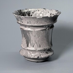 May include: A metallic silver vase with a flared, uneven rim and a textured, rippled surface. The vessel features a wide opening that tapers towards a narrow base, displaying a reflective, molten-like finish with dark grey and silver highlights.