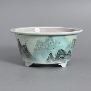 May include: A light blue ceramic planter bowl with a wide rim and three small feet. The exterior features a hand-painted landscape scene in black ink, depicting jagged mountains and several flying cranes against a soft, pale blue background.