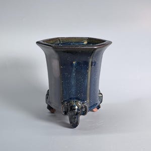 Op de afbeelding: Een zeshoekige keramische plantenbak met een glanzend donkerblauw glazuur met witte spikkels. De pot heeft een uitlopende rand en rust op drie gebeeldhouwde voetjes in de vorm van olifantenkoppen, elk met zichtbare slagtanden en oren in blauw.