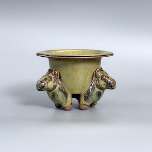 May include: A ceramic planter with a wide, flat rim and a speckled olive-green glaze. The base is supported by three sculpted figures resembling stylized horses or animals, featuring brown and green tones with a glossy, smooth finish.