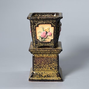 Op de afbeelding: Een vierkante keramische plantenbak met een donkerbruin en goud gespikkeld glazuur, voorzien van een verhoogd, lichtgekleurd paneel aan elke zijde met een reliëf van roze en gele bloemen en groene bladeren, rustend op een bijpassende voet.
