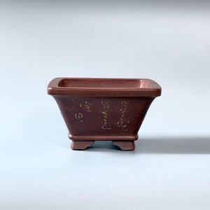 Peut inclure: Un pot à bonsaï rectangulaire en céramique brun-rouge foncé, doté d'un bord large et plat ainsi que de quatre petits pieds robustes. La face avant présente des caractères calligraphiques clairs gravés sur la surface mate et lisse.