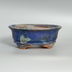 Puede incluir: Una maceta de bonsái de cerámica azul con un acabado moteado. El recipiente de forma ovalada tiene un borde ancho y se apoya en tres pequeños pies decorativos. El interior y el exterior de la maceta son de color azul oscuro, con toques de verde y marrón.