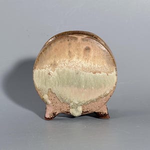 Könnte beinhalten: Ein kreisförmiges Keramikgefäß mit drei kleinen Füßen, das mit einer reaktiven Glasur in Beige-, Hellbraun- und gedämpften Grüntönen versehen ist. Die Oberfläche weist ein gesprenkeltes, strukturiertes Aussehen mit einem horizontalen Farbverlauf auf.