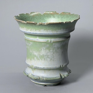 May include: A ceramic vase featuring a flared, scalloped rim and a tapered body with horizontal ridges. The surface displays a gradient of soft sage green and muted white tones, with subtle blue-grey accents along the textured edges and base.