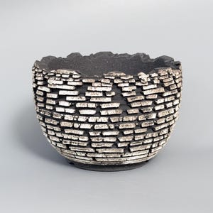 May include: A round ceramic bowl with a dark black interior and a textured exterior featuring a layered, brick-like pattern in shades of black and metallic silver. The rim has an uneven, jagged edge, and the surface has a rough, weathered appearance.