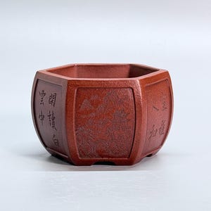 May include: A hexagonal terracotta-colored ceramic planter featuring intricate relief carvings on its exterior panels. The central panel displays a detailed landscape with mountains and a pavilion, while the side panels contain engraved Chinese calligraphy.