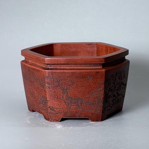 Puede incluir: Una maceta hexagonal de cerámica de color marrón rojizo con una escena de paisaje grabada que muestra un ciervo en un bosque. El recipiente tiene un borde ancho, una superficie texturizada y pequeños pies en la base con detalles grabados.