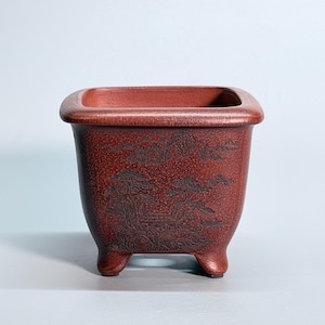 May include: A square, reddish-brown ceramic planter with a wide, rounded rim and four short, sturdy feet. The exterior surface features a detailed, dark-toned engraved landscape scene depicting mountains, pine trees, and a small structure.