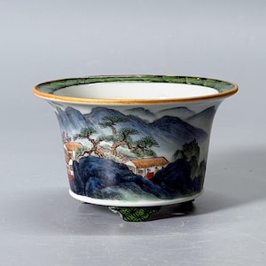 May include: A white ceramic planter with a flared rim and three small feet. The exterior features a hand-painted landscape scene with blue mountains, green pine trees, and small buildings. The rim has a green patterned border with a gold edge.