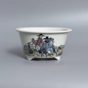 Puede incluir: Un cuenco de cerámica blanca con borde acampanado y tres pies pequeños. El exterior presenta una escena pintada a mano que muestra figuras con vestimenta tradicional, incluida una persona montando un burro oscuro, en tonos azules, rojos y marrones.