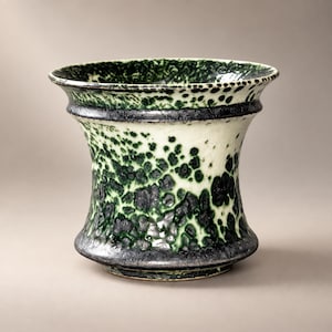 以下が含まれることがあります： 広がった縁と先細りの胴体を持つ陶器製の花瓶。クリームがかったオフホワイトのベースに、ダークフォレストグリーンとブラックの釉薬が斑点状に施されています。表面は質感があり、光沢のある仕上げで、上部は広く開いています。