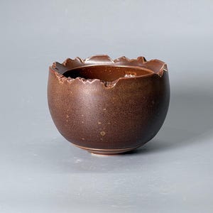 Może przedstawiać: Okrągła, ciemnobrązowa miska ceramiczna o nakrapianym, matowym wykończeniu i poszarpanym, nierównym brzegu. Miska posiada głębokie, gładkie wnętrze oraz małą, okrągłą podstawę, prezentując teksturowany, ziemisty wygląd na całej powierzchni.