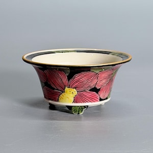 May include: A ceramic bowl with a flared rim, featuring a black exterior decorated with large pink flowers and a small yellow cat nestled among the petals. The bowl has a cream-colored interior and rests on three small feet with a gold-toned rim.