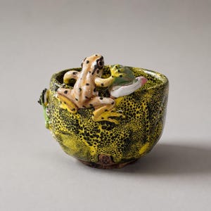 Puede incluir: Un cuenco de cerámica con esmalte moteado en amarillo y negro, que presenta una rana esculpida de color marrón claro con manchas oscuras posada en el borde, junto a una figura de rana verde más pequeña en el exterior.