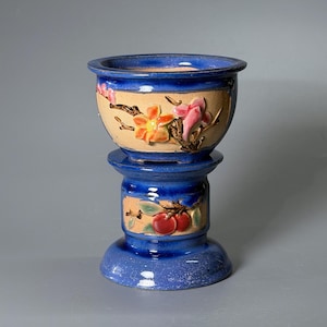 Puede incluir: Una maceta de cerámica de dos piezas con acabado esmaltado en azul intenso y paneles beige. El cuenco superior muestra flores en relieve de color naranja y rosa, mientras que la base cilíndrica presenta frutas rojas y hojas verdes.