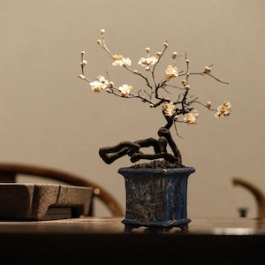 Könnte beinhalten: Ein dekorativer Bonsai-Baum mit dunklen, gedrehten Ästen und kleinen, cremeweißen Blüten. Der Baum ist in einen quadratischen, tiefblauen Keramiktopf gepflanzt, der an den Seiten mit detaillierten Reliefs verziert ist und auf vier kleinen Füßen steht.