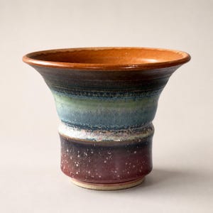 May include: A ceramic vase with a wide, flared rim and a tapered body. The exterior features a multi-coloured glaze with bands of deep red at the base, transitioning into mottled blue and green tones, and a warm, unglazed terracotta rim.