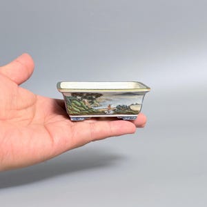 Op de afbeelding: Een rechthoekige keramische bonsaipot met een handgeschilderd landschap met groene heuvels, bomen en een klein figuurtje op een pad. De pot heeft een witte binnenkant, een goudkleurige rand en vier kleine voetjes met blauw-witte patronen.